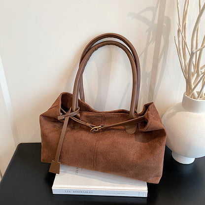 Brown suede handbag on a stack of books with a white vase and decorative branches in the background.